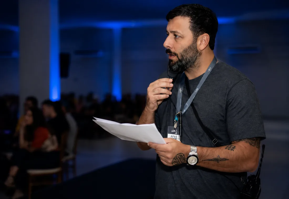 Gabriel Penello dirigindo um evento, rádio e crachá no pescoço, prancheta na mão